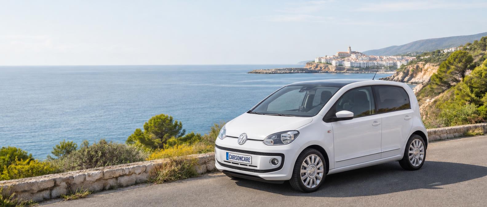 A white Volkswagen Up! with a CARSONCARE number plate on a sunlit Spanish coastal road